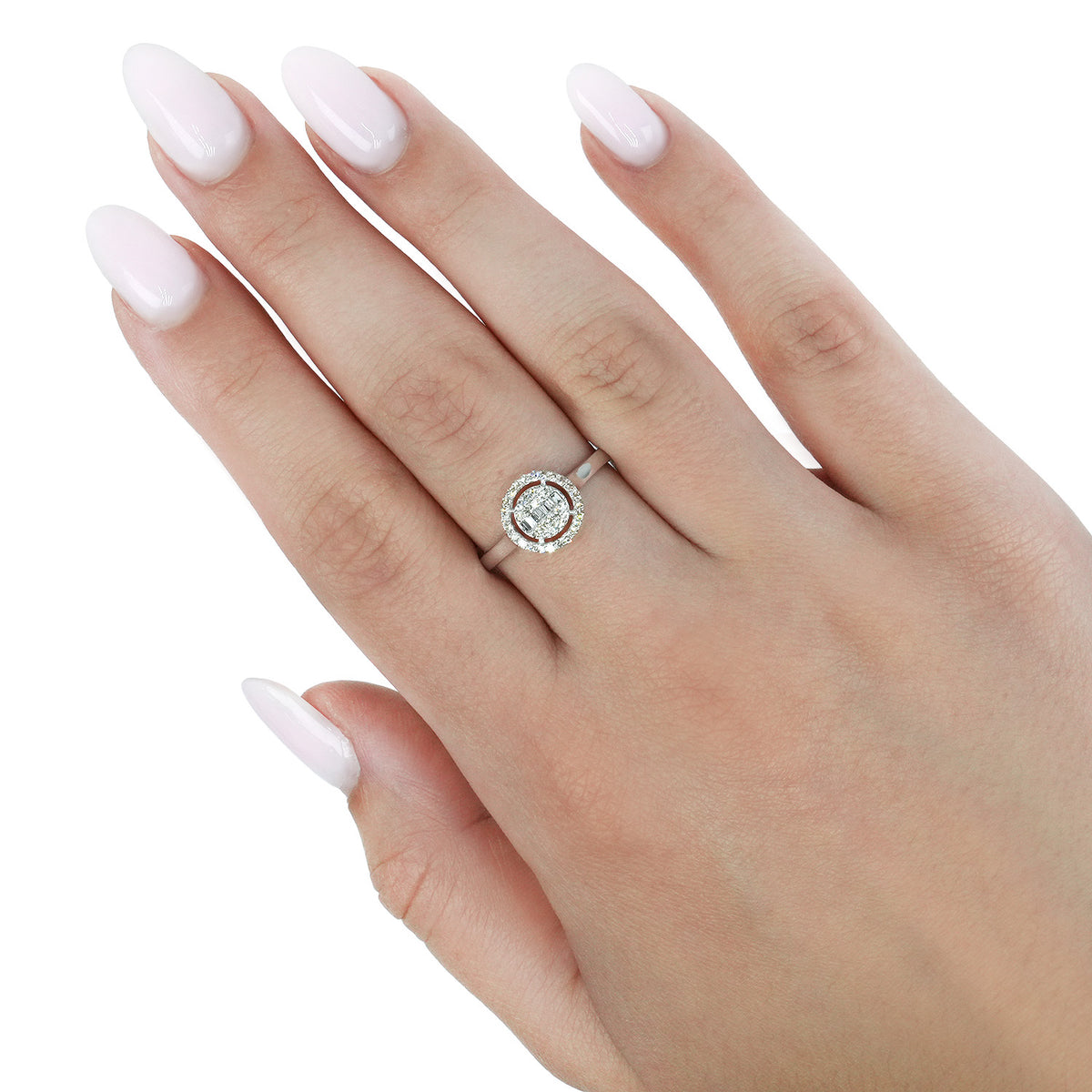 A silver ring with a round halo of small clear stones worn on a hand with pale pink manicured nails, set against a plain white background. [lifestyle]