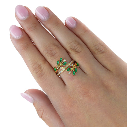 A close-up of a hand with light pink manicured nails wearing a gold wrap ring decorated with green leaf-shaped enamel and small clear stones. [lifestyle]