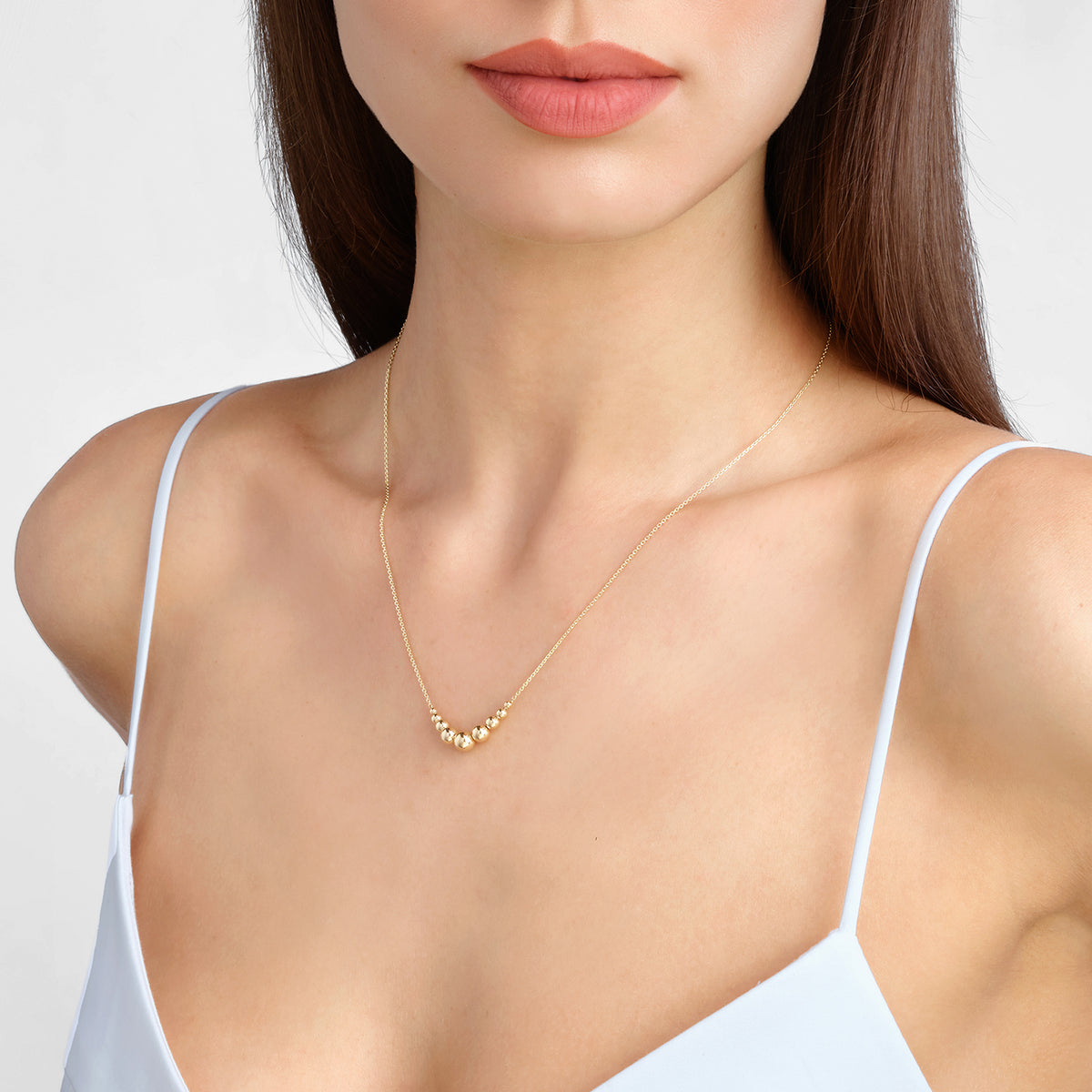 A close-up of a woman wearing a delicate gold necklace with a small cluster of pearl-like beads at the center, paired with a white spaghetti-strap top against a light gray background. [lifestyle]