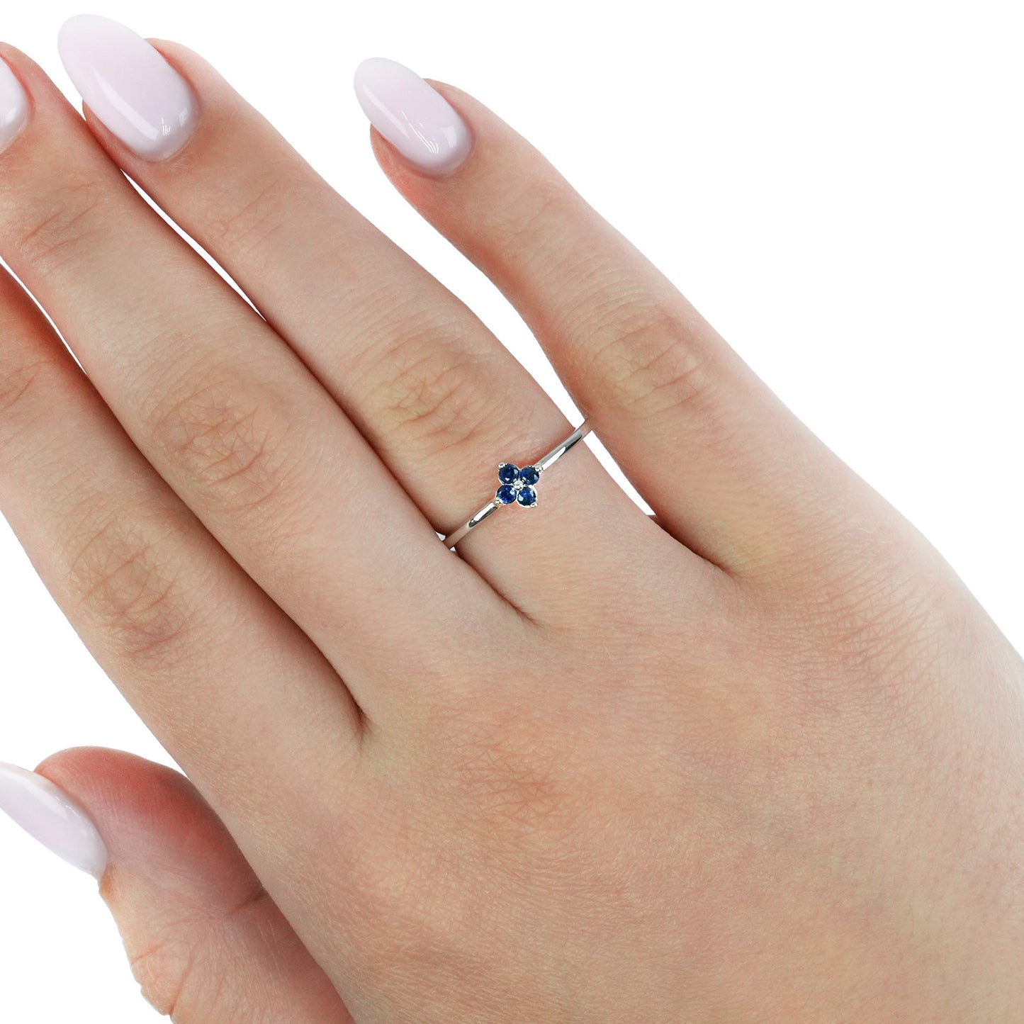 A silver ring with a small four-stone floral cluster featuring blue and white stones, shown worn on a hand with pale pink manicured nails against a white background. [lifestyle]