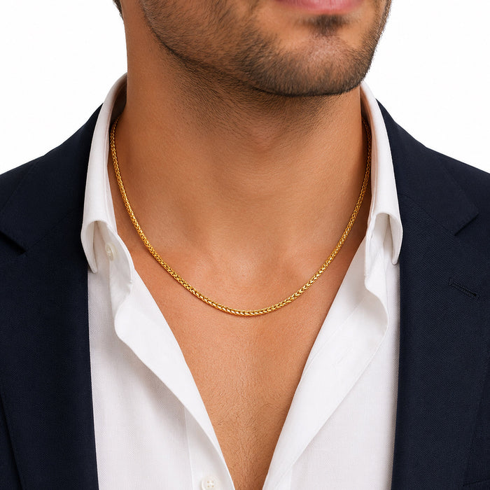 A close-up of a man wearing a delicate gold chain necklace over a white collared shirt and dark navy blazer, against a plain white background. [lifestyle]