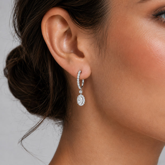 A close-up of a woman's ear wearing a silver-toned drop hoop earring with a teardrop-shaped clear gemstone and small pavé accents. [lifestyle]