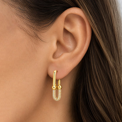 A close-up of a model's ear wearing a gold huggie hoop earring with a dangling U-shaped charm adorned with small white pearls. [lifestyle]