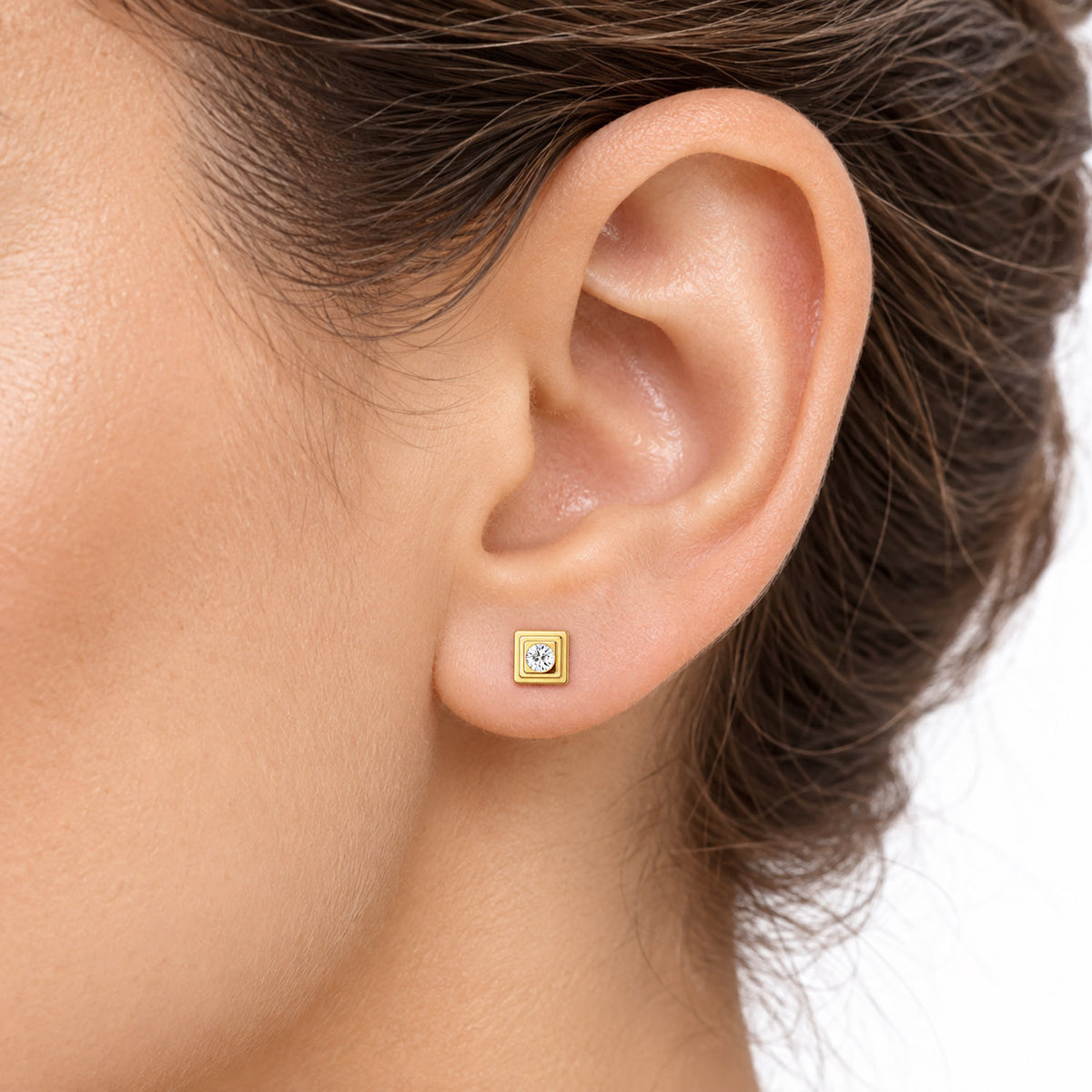 A close-up of a person's ear wearing a small square gold stud earring with a clear gemstone, set against a clean white background. [lifestyle]