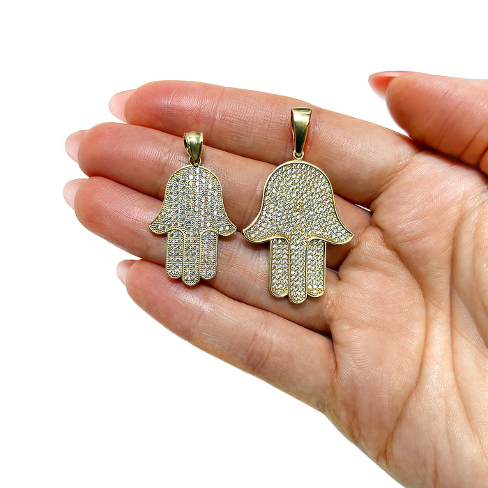 Two gold Hamsa hand pendants covered in clear pavé stones, held in a person's hand against a white background. [lifestyle]